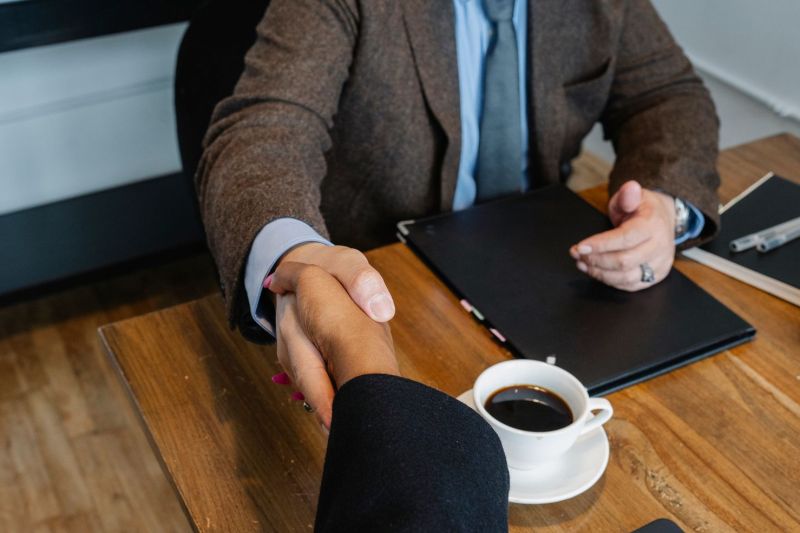 Two people shaking hands across a table holding documents and a coffee cup.